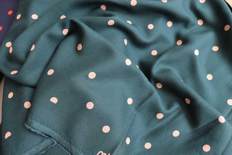 Puede incluir: Una tela de color verde azulado con un patr&oacute;n repetido de lunares rosa claro. La tela est&aacute; doblada y drapeada, mostrando su textura y la forma en que captura la luz. El efecto general es suave y fluido.