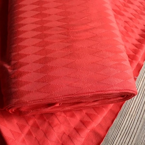 Könnte beinhalten: Ein aufgerolltes Stück roter Stoff mit einem sich wiederholenden Rautenmuster. Der Stoff ist auf sich selbst gefaltet, wodurch die Textur und das Design sichtbar werden. Der Stoff liegt auf einer Holzoberfläche.