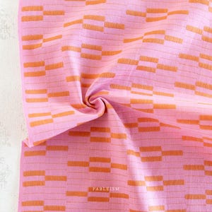 Peut inclure: Tissu rose avec un motif rectangulaire orange répété. Le tissu est plié, créant un tourbillon au centre. Le mot "FABLEISM" est imprimé en blanc en bas à droite.