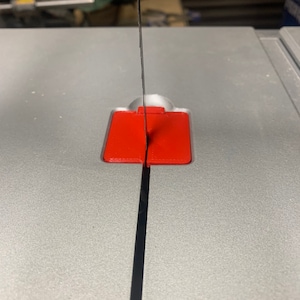 May include: A close-up of a black saw blade cutting through a red plastic guide on a gray surface.