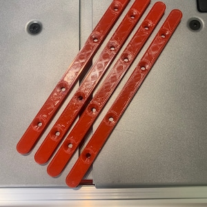 May include: Four red, elongated plastic pieces with multiple circular holes, arranged diagonally on a gray surface. The pieces appear to be components of a larger item. The image includes a ruler with centimeter markings.