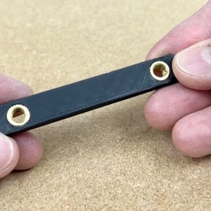 May include: A small, black rectangular object with two brass-colored circular inserts. The object is held between two hands. The item appears to be a small hardware component.