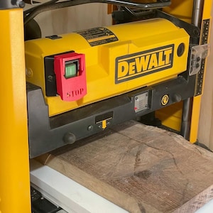 May include: A yellow and black DEWALT planer with a red stop button. A piece of wood is being fed into the planer.