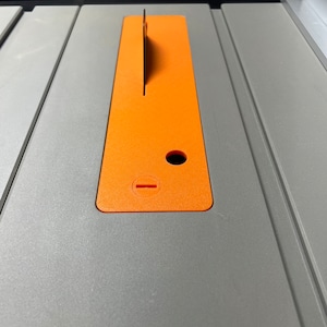 May include: A close-up of a table saw with an orange plastic insert. The insert has a slot for a blade and a hole for a dust collection port.