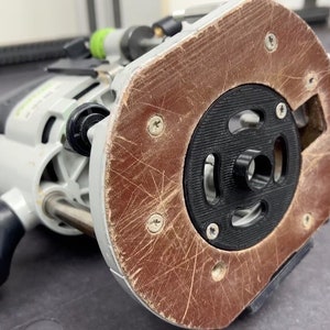 May include: A close-up of a gray and black router with a brown base plate. The base plate has a black center with a hole in the middle. The router is on a gray surface.