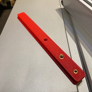 May include: A red plastic ruler with three brass screws. The ruler has a textured surface and a hole in the center.