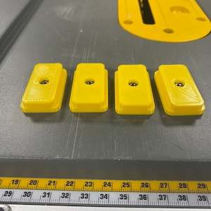 May include: Four yellow plastic rectangular blocks with a small metal screw in the centre of each block. The blocks are arranged in a row on a grey surface. A ruler with black numbers is visible at the bottom of the image.
