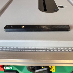 May include: A black plastic table saw accessory with three holes and a brass screw. The accessory is sitting on a gray table surface with a ruler marked in inches.