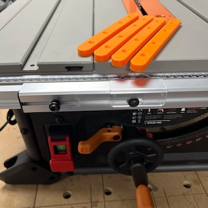 May include: A black table saw with a grey table top and orange plastic fence extensions. The saw has a red lever and a black knob. The table saw is on a wooden surface with holes.
