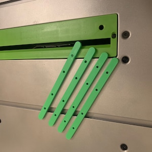May include: Four green plastic strips with holes, possibly for a table saw or other woodworking tool.