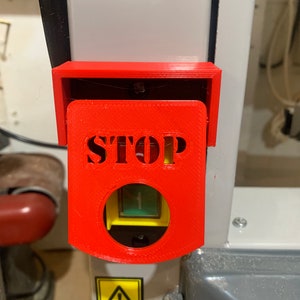 May include: A bright red STOP button cover with the word "STOP" cut out. The cover is mounted on a machine with a yellow and black warning label. The button itself is yellow and green.