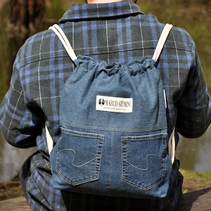 Op de afbeelding: Een blauw en zwart geruit overhemd met een denim rugzak met trekkoord. De rugzak heeft een wit label met de tekst "MARCO GEMINI".