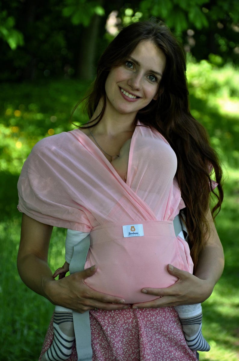 May include: A woman wearing a pink wrap-style baby carrier with a white and blue striped pattern. The carrier has a label that says "Sunbaby" on it. The woman is holding a baby in the carrier.