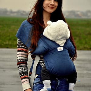 May include: A woman wearing a blue and white striped sweater and blue jeans is carrying a baby in a blue baby carrier. The baby is wearing a white hooded jacket.