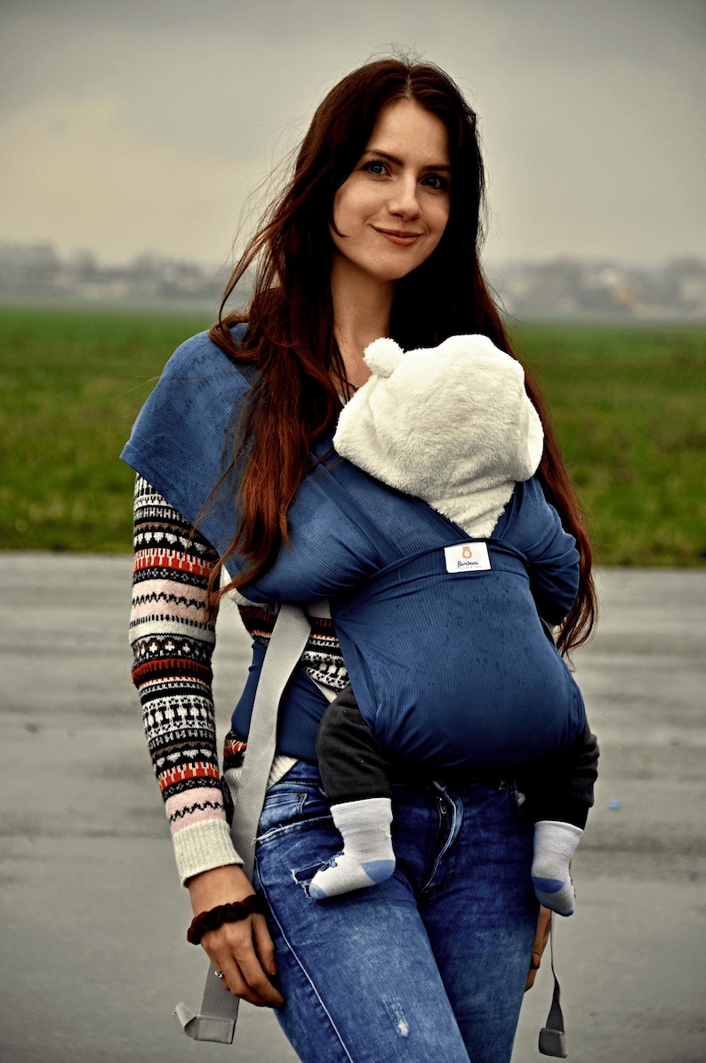May include: A woman wearing a blue and white striped sweater and blue jeans is carrying a baby in a blue baby carrier. The baby is wearing a white hooded jacket.