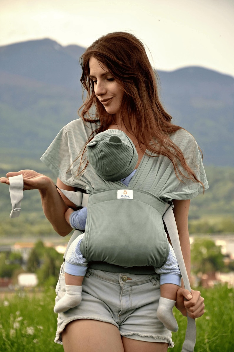 May include: A woman wearing a light blue shirt and denim shorts is carrying a baby in a green baby carrier. The carrier has a white strap and a small logo that says "Bamboo".