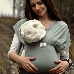 May include: A woman wearing a gray baby carrier with a white plush toy inside. The carrier has a small brown label with the word "Bambino" on it.