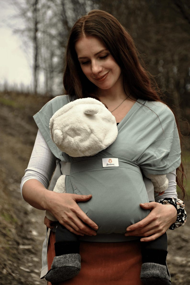 May include: A woman wearing a gray baby carrier with a white plush toy inside. The carrier has a small brown label with the word "Bambino" on it.