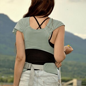 May include: A woman wearing a light green and black baby carrier with a gray belt. She is wearing denim shorts and a black tank top. The woman is standing outdoors with a mountain range in the background.