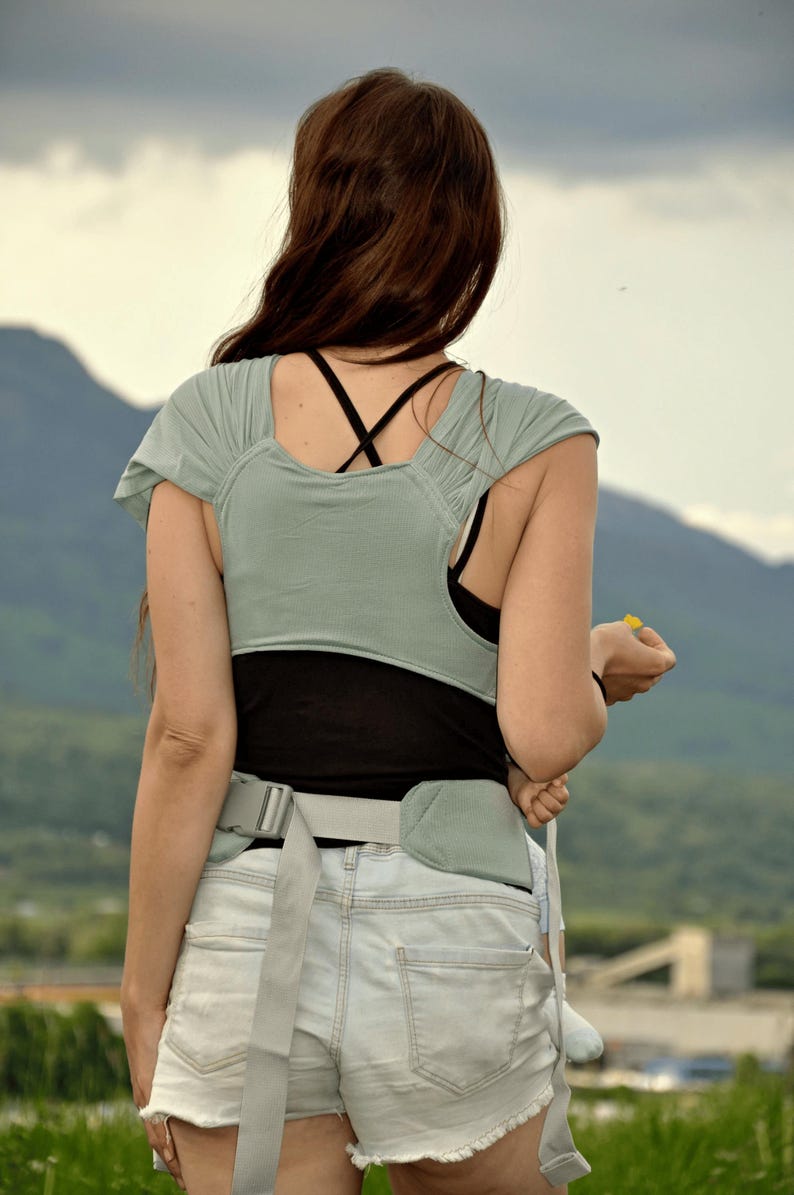 May include: A woman wearing a light green and black baby carrier with a gray belt. She is wearing denim shorts and a black tank top. The woman is standing outdoors with a mountain range in the background.