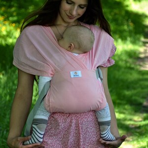 May include: A woman wearing a pink baby wrap carrier with a baby sleeping inside. The baby is wearing a grey and white striped shirt and socks. The woman is wearing a pink floral dress.