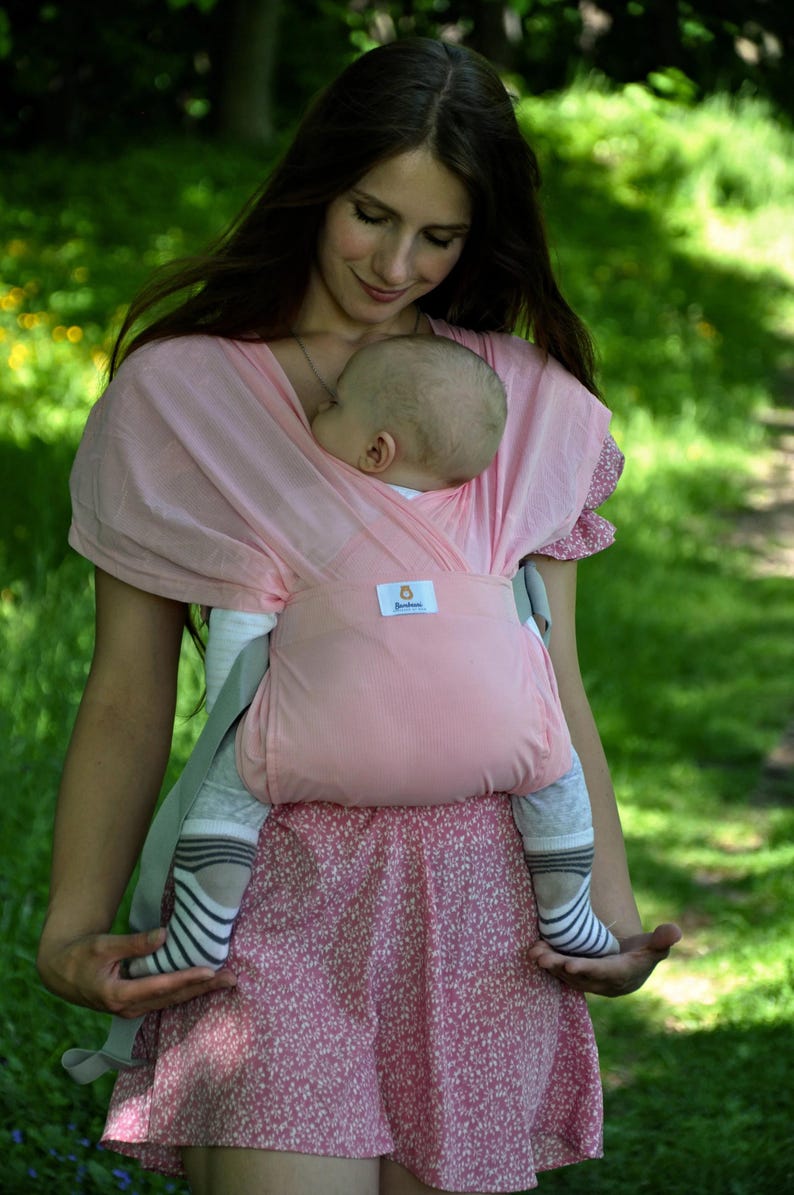 May include: A woman wearing a pink baby wrap carrier with a baby sleeping inside. The baby is wearing a grey and white striped shirt and socks. The woman is wearing a pink floral dress.