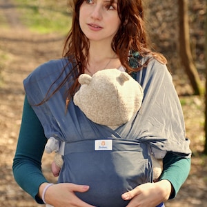 May include: A woman wearing a blue baby wrap carrier with a baby inside. The baby is wearing a blue and white striped onesie and a brown knitted hat. The woman is holding the baby close to her chest. The baby wrap carrier has a label that says "Soothe Baby".