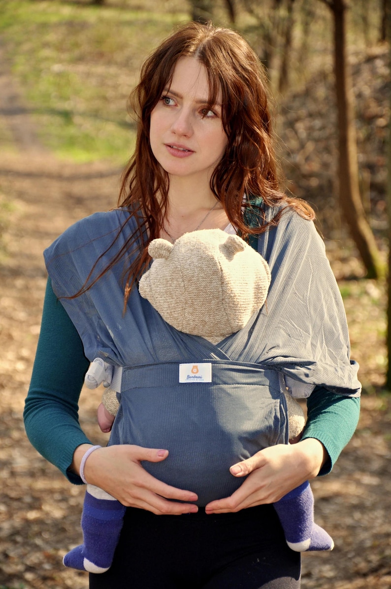 May include: A woman wearing a blue baby wrap carrier with a baby inside. The baby is wearing a blue and white striped onesie and a brown knitted hat. The woman is holding the baby close to her chest. The baby wrap carrier has a label that says "Soothe Baby".