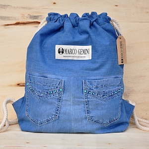 Op de afbeelding: Blauwe denim trekkoordtas met twee voorzakken versierd met turquoise strass steentjes. De tas heeft een label met de tekst "MARCO GEMINI" en een kleiner label met de tekst "MARCO GEMINI".