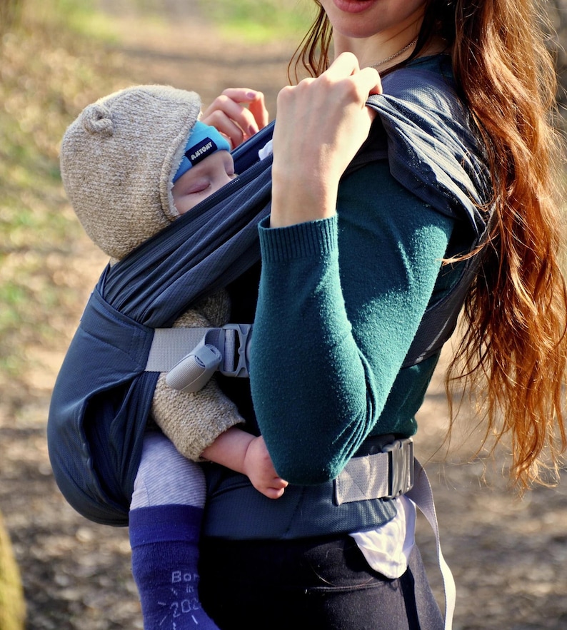 May include: A woman wearing a teal shirt carries a sleeping baby in a dark blue baby carrier. The baby is wearing a beige knitted hat with a blue tag that says "PLATINUM".