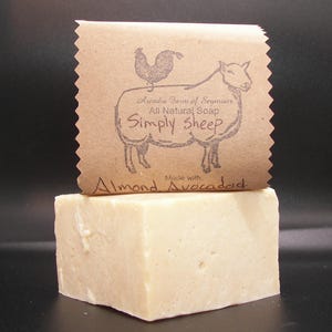 Puede incluir: Una pastilla de jabón rectangular de color beige claro con una etiqueta de papel marrón. La etiqueta presenta una ilustración de oveja con el texto "Simply Sheep" y "Almond Avocado". El jabón es de Arcadia Farm of Seymour.