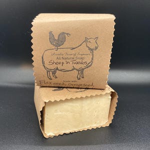 Pode incluir: Uma barra de sabonete natural, embrulhada em papel castanho. O papel é impresso com uma ilustração de ovelha e galo, e o texto "Sheep in Tuxedos". Contém sementes de linhaça e sementes de uva.