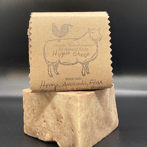 Op de afbeelding: Een natuurlijke zeep in blok met een bruin papieren etiket waarop staat "Arcadia Farm of Seymour Natuurlijke Zeep Hippie Sheep Gemaakt met: Hennep, Avocado, Lijnzaad Olie & Shea Butter".