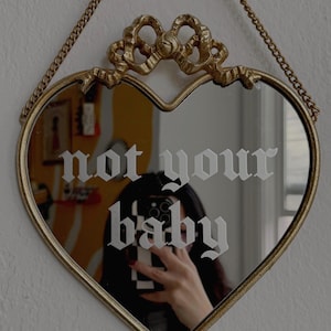 May include: Heart-shaped mirror with a gold frame and chain. The mirror has the words "not your baby" etched in white. The top of the mirror features a decorative bow design.