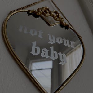 May include: Heart-shaped mirror with a gold-colored frame and chain. The mirror reflects a window and the text "not your baby" in a gothic font. The top of the frame has a decorative gold accent.