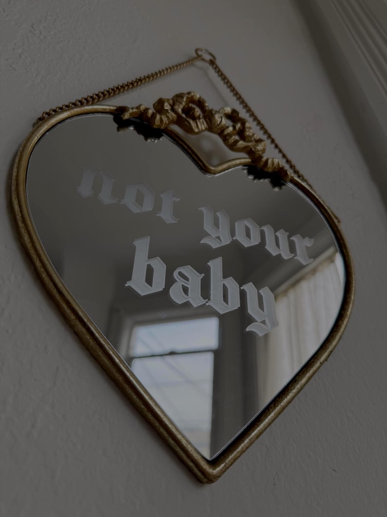 May include: Heart-shaped mirror with a gold-colored frame and chain. The mirror reflects a window and the text "not your baby" in a gothic font. The top of the frame has a decorative gold accent.