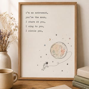 Könnte beinhalten: Ein gerahmter Kunstdruck mit einer Strichzeichnung eines Astronauten, der mit einem Aquarellmond verbunden ist, umgeben von kleinen Sternen. Der Text lautet: i'm an astronaut, you're the moon, I stare at you, I sing to you, I circle you, in Schreibmaschinenschrift.