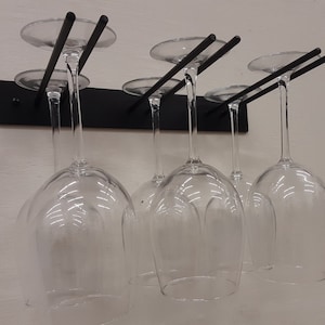 May include: A black metal wine glass rack mounted on a wall, holding six clear wine glasses. The rack has a minimalist design with horizontal bars to support the glasses. The glasses are suspended upside down, showcasing their elegant shape.