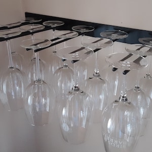 May include: A black metal wine glass rack mounted on a white wall, holding multiple clear wine glasses. The glasses are suspended upside down, showcasing their stems and bowls. The rack is designed to hold the glasses securely.