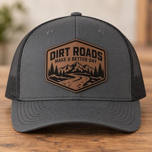 May include: A charcoal gray trucker hat with a black mesh back and a brown leather patch on the front. The patch features a black graphic of a winding dirt road leading toward mountains and pine trees, with the text DIRT ROADS MAKE A BETTER DAY.
