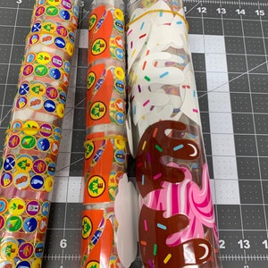May include: Three rolls of stickers wrapped in clear plastic. The first roll has colorful stickers with images of camping gear, the second roll has orange stickers with images of trees and the third roll has white stickers with images of ice cream cones.