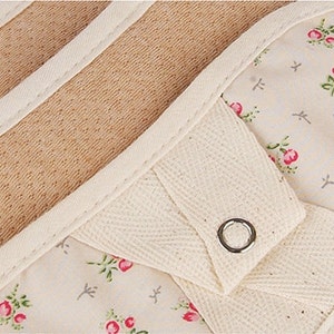 May include: A pair of beige oven mitts with a floral print and a white cotton strap with a metal button. The mitts are lined with a brown, textured material.