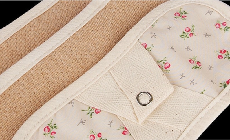 May include: A pair of beige oven mitts with a floral print and a white cotton strap with a metal button. The mitts are lined with a brown, textured material.