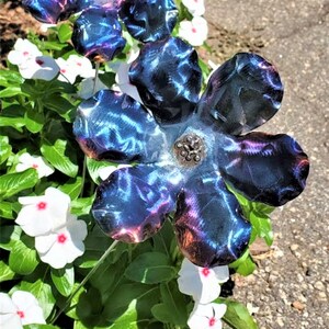 May include: Two metal flowers with a blue and purple iridescent finish. The flowers are made of metal and have a textured surface. The flowers are in a garden setting with white flowers in the background.
