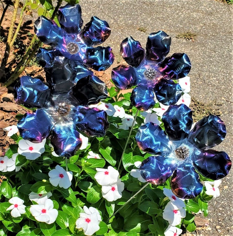 May include: Four metal flowers with iridescent blue and purple coloring. The flowers are arranged in a cluster and are placed in a bed of white flowers.