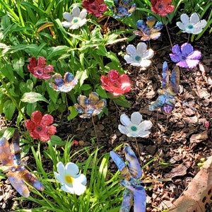 May include: A collection of colorful ceramic flowers and dragonflies, with iridescent finishes, arranged in a garden setting. The flowers are in shades of red, white, and purple, while the dragonflies are blue and brown.