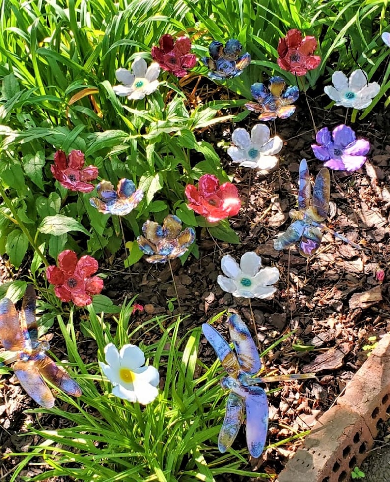 May include: A collection of colorful ceramic flowers and dragonflies, with iridescent finishes, arranged in a garden setting. The flowers are in shades of red, white, and purple, while the dragonflies are blue and brown.