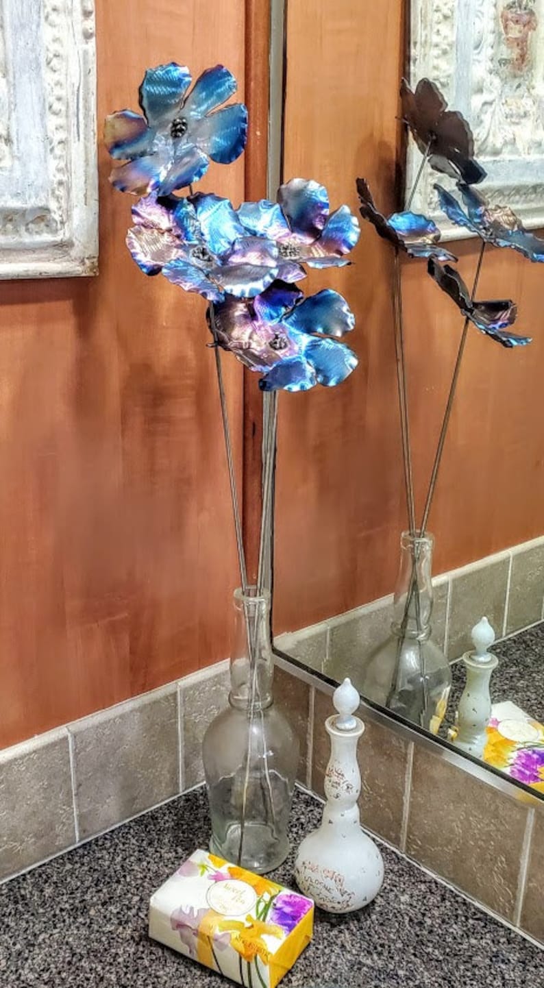 May include: A metal flower arrangement with blue and purple flowers in a clear glass vase. The flowers are on a black countertop with a white and yellow floral patterned soap bar.