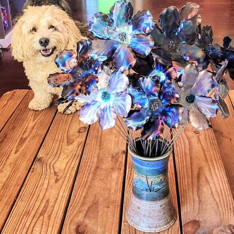 May include: A bouquet of iridescent blue and purple metal flowers in a rustic brown and blue ceramic vase. The flowers are arranged on a wooden table.
