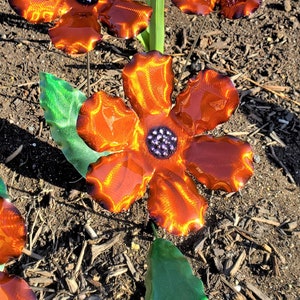 May include: Three metal flowers with orange petals and purple centers. The flowers are on green stems with green leaves. The flowers are sitting on a bed of brown mulch.